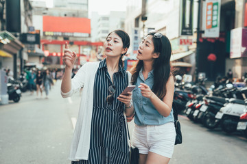 Fototapeta premium Friends having fun outdoors gathered together looking at smart phone on holiday. two young asian girls traveler holding cellphone pointing searching direction on street with scooter taiwan taipei