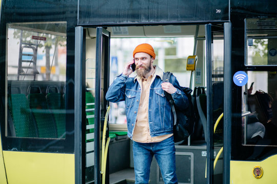 Portrait Of Happy Young Hipster American Travel Man In Blue Denim Jacket And Orange Hat With Bag Getting Off Bus Talking On Phone On The Stop.