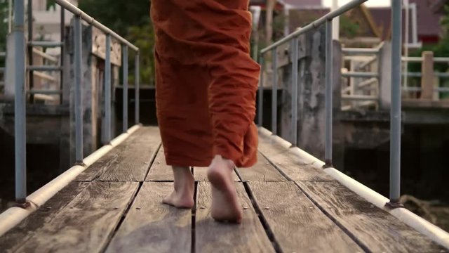The monks are walking down to the waterfront using a wooden bridge. Slow motion.