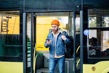 Portrait of happy young hipster american travel man holds smartphone dressed in blue denim jacket and orange hat with bag getting off bus talking on phone on the stop.