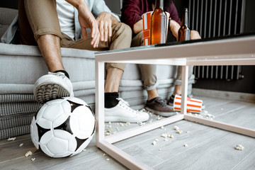 Friends watching football match. View on their legs with pop cornes and ball on the floor