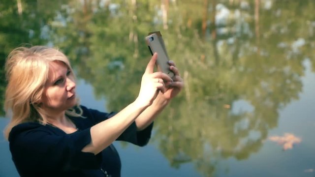 portrait young blonde woman taking selfie with a mobile phone on a walk against the backdrop of a pond city park walk