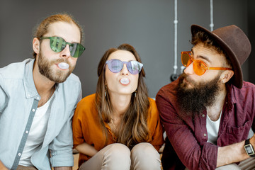 Young friends in colorful glasses having fun with gum bubbles sitting together at home