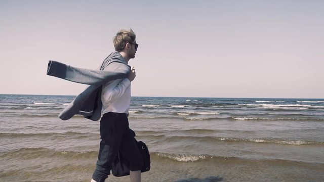 Slow motion footage of young business man walking in beach with his jacket in one hand and laptop PC in other. Sea with waves and blue sky in background.