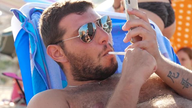 A man relaxes on a beach while having a cigarette and checking his mobile phone