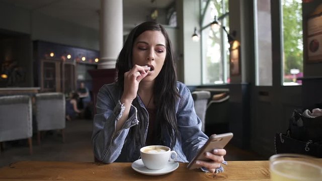Attractive hispanic brunette sitting at the table, eating a cookie and having a coffee while checking her phone