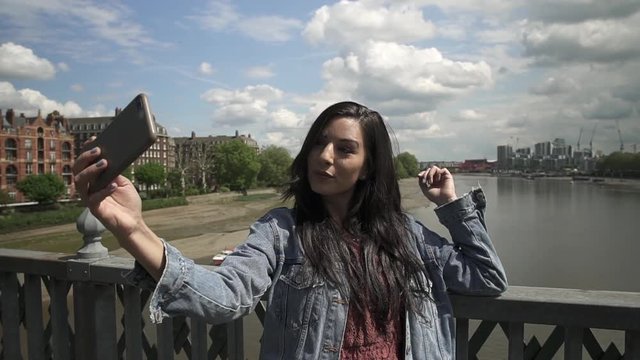 Brunette Latina tourist taking a selfie, posing doing victory sign with her hand, while standing on the railing of a bridge in London