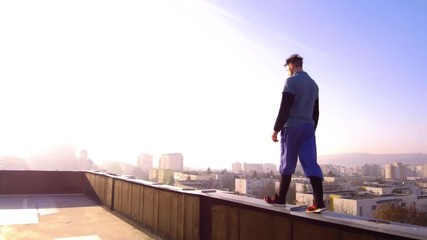 Man walking on the edge of a building rooftop toward bright sunlight in the distance. Buildings of the city far below and on the horizon. TRACKING. - Powered by Adobe