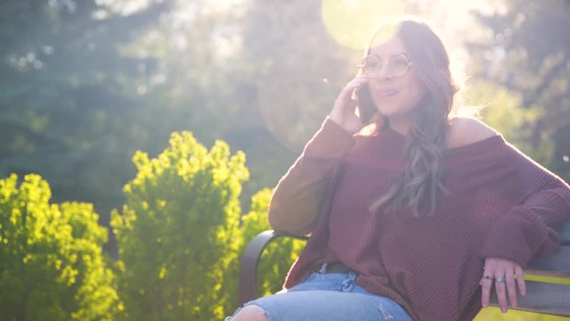 A Young Woman Sitting On A Park Bench Talking On The Phone While Laughing. Wide Shot.