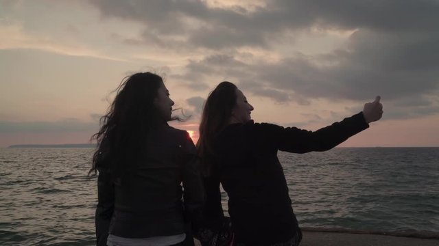 Two girls are taking selfie of themselves near the sea at the sunset