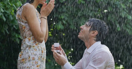 Portrait of young handsome man is making a proposal of marriage to his fiancee under the rain on a background of green trees.