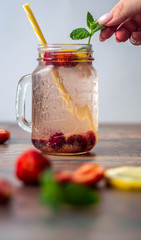Woman decorates a cocktail in a glass beaker with fresh mint leaves on a white background. In the foreground are strawberries, lemon and mint.