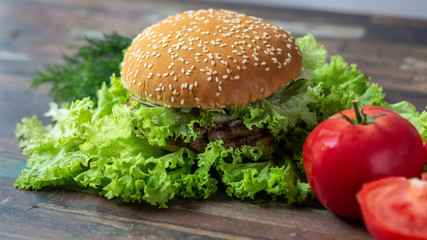 Juicy, tasty burger lies on lettuce leaves with red tomato, dill on wooden background