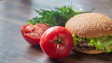 A juicy, big, red tomato in the foreground lies on a wooden background with dill and a tasty big  burger