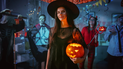 Halloween Costume Party: Gorgeous Seductive Witch Wearing Dress Holds Burning Pumpkin. Background: Beautiful Devil, Scary Death, Count Dracula, Zombie Dancing in the Decorated Room