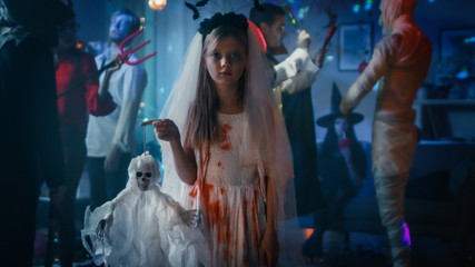 Halloween Costume Party: Little Girl in a Bloody White Bride Dress Holding Scary Doll. In the Background Group of Monsters Dancing and Having Fun in Decorated Room with Disco Ball Light Shot