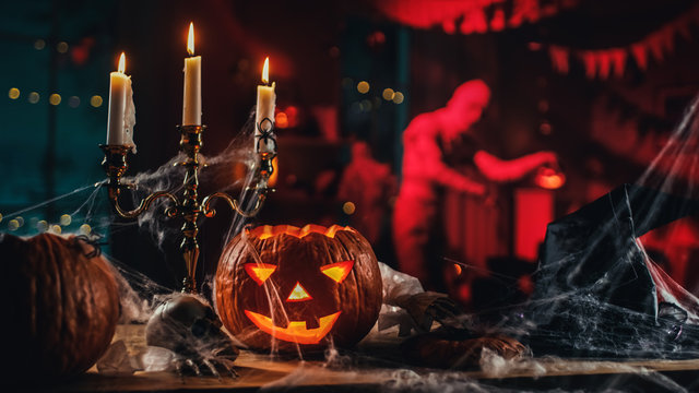 Halloween Still Life Colorful Theme: Scary Decorated Dark Room with Table Covered in Spider Webs, Burning Pumpkin, Candlestick, Witch's Hat and Skeleton. In Background Silhouette of Monster Walking By