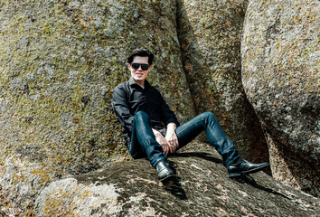Naklejka na ściany i meble young handsome guy sitting on a big stone in black glasses and a black shirt and jeans in summer