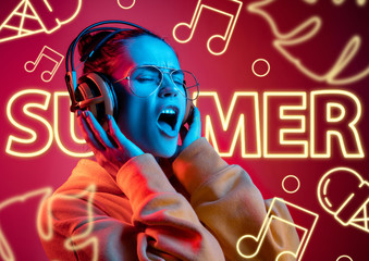 Female model in trendy neon light on red background. Modern design. Beautiful young woman listen to music in headphones and singing. Concept of facial expression, summer, vacation, music, open-air.