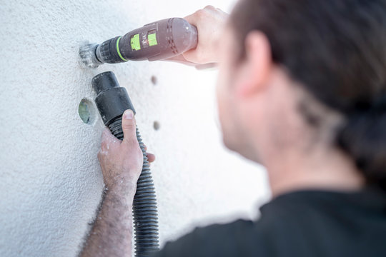 Handwerker bohrt mit dem Akkuschrauber Loch in die Wand und fr&auml;st es mit dem Putzfr&auml;ser gr&ouml;&szlig;er und sein Lehrling hilft ihm indem er den Staub absaugt