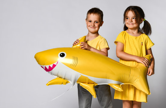 Positive young children hold a balloon in the shape of a yellow shark fish, celebrate the holiday, smiling widely, stand on a light background, are in a good mood