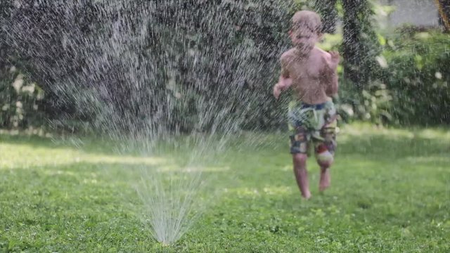Slow Motion of Young Boy Child Running Over Sprinkler With Sprinking Water in Yard on Hot Summer Day