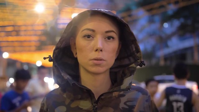 Slow motion view of woman standing outside wearing a camo jacket with hood over her head. Lady unzips camouflage jacket to reveal black shirt under and takes hood off her head.