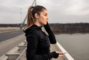 Portrait of young woman in black sports outfit resting after running on the bridge in the city.