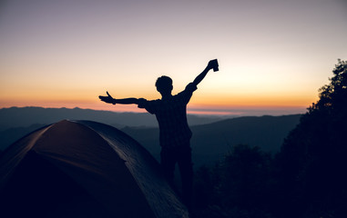 Happy hiker man holding coffee cup near camping tent on mountains at sunset background. travel concept.