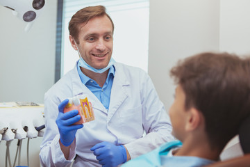 Mature cheerful male dentist educationg his young patient, showing tooth model, explaining teeth care. Professional dentist talking to a little boy