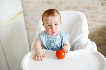 Fototapeta premium Baby eating vegetables. red tomato in little girl hand in sunny kitchen. Healthy nutrition for kids. Snack or breakfast for young child.