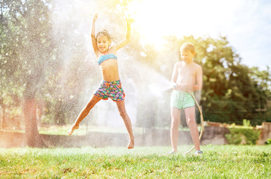 Happy little girl jumps under water, when brother pours her from garden hose. Hot summer days activity.