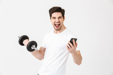 Emotional happy excited young man posing isolated over white wall holding dumbbell make exercise using mobile phone.