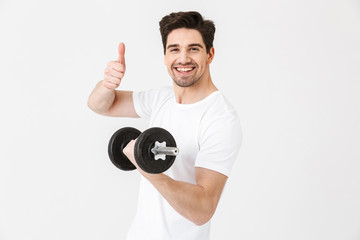 Emotional happy excited young man posing isolated over white wall holding dumbbell make exercise make thumbs up gesture.