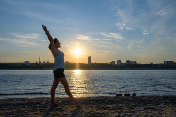 Fototapeta na wymiar The girl is engaged in yoga, gymnastics, doing exercises by the river against the sunset and the city. The warm rays of the setting sun, blue sky and a romantic tint.