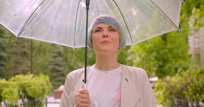 Closeup portrait of young cute caucasian female with dyed hair holding an umbrella walking towards camera outdoors in the park