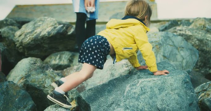 Little toddler climbing rocks on beach with mother and grandfather watching