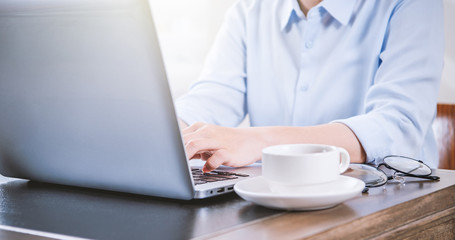 Business concept. Woman in blue shirt typing on computer with coffee on office table, backlighting, sun glare effect, close up, side view, copy space