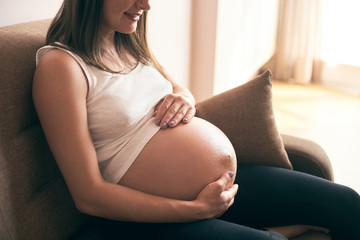 Cropped view of pregnant woman in white shirt sitting on sofa at home, embracing belly and smiling. Happy mother enjoying pregnancy and expecting little baby. Concept of happiness.