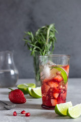 Homemade strawberry lemonade in glass with fresh strawberries, rosemary, pomegranate and lime over light grey stone table. Refreshing summer drink. Cocktail bar background concept. Copy space.