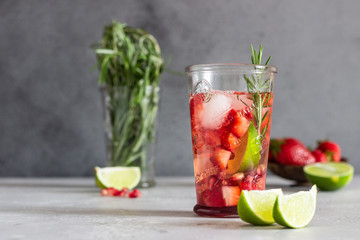 Homemade strawberry lemonade in glass with fresh strawberries, rosemary, pomegranate and lime over light grey stone table. Refreshing summer drink. Cocktail bar background concept. Copy space.