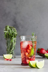 Homemade strawberry lemonade in glass with fresh strawberries, rosemary, pomegranate and lime over light grey stone table. Refreshing summer drink. Cocktail bar background concept. Copy space.
