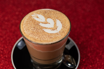A close up view of a latte art flower on top of a coffee in a drinking glass against a red sparkling background