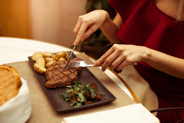 Focus on the meat where the fork and knife. Beautiful female hands in a restaurant at the table cut beef striploin steak.