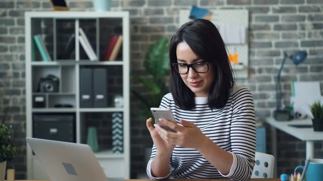 Beautiful woman in glasses is using smartphone touching screen smiling sitting at desk in office room alone enjoying gadget. People, workplace and devices concept.
