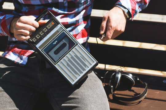 Hipster fancy man in plaid shirt holding in hand and sitting on bench with retro vintage old 80s , 90s style portable radio audio cassette recorder player boombox with audio tape cassette inside 