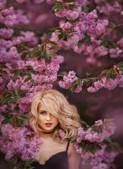 Fototapeta premium Sexy blonde looks with a piercing gaze ,among the flowering branches of Sakura ,the wind blows off her hair