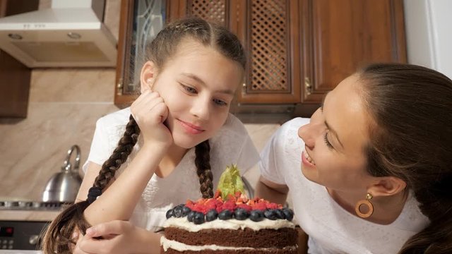 Portrat of mom and daughter are sniffing cooked homemade chocolate cream cake with blueberries, raspberries and physalis in kitchen at home. Teen girl is taking berry from the cake and eating it.