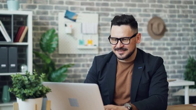 Portrait of attractive entrepreneur joyful guy in jacket and glasses using laptop then looking at camera smiling. Business people and modern technology concept.