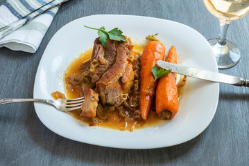 Roasted pork with carrots and onion sauce in a plate close-up and a glass of white wine on a wooden dark table, top view - French, European cuisine